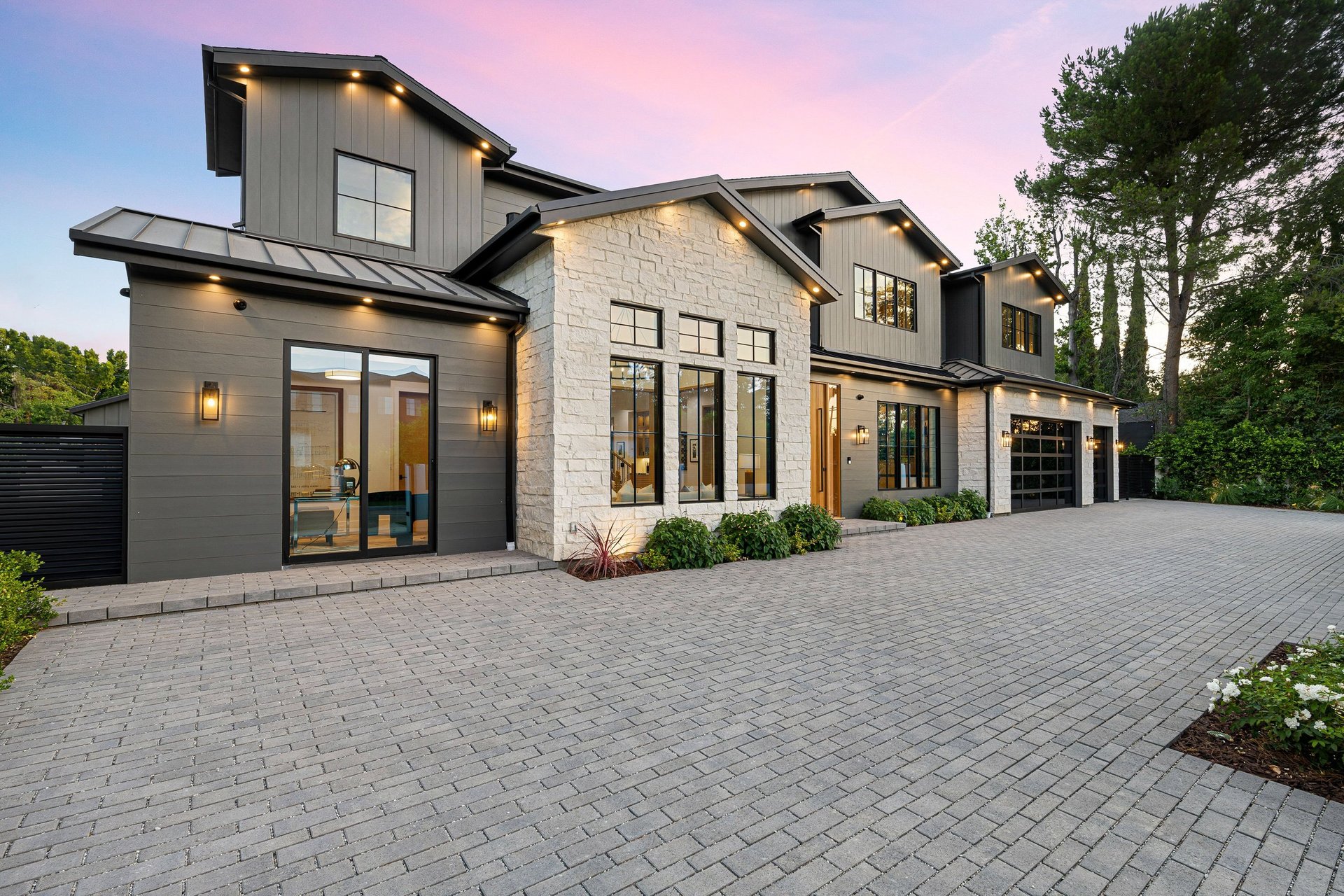 Modern luxury home exterior with large windows, stone facade, and spacious driveway at sunset