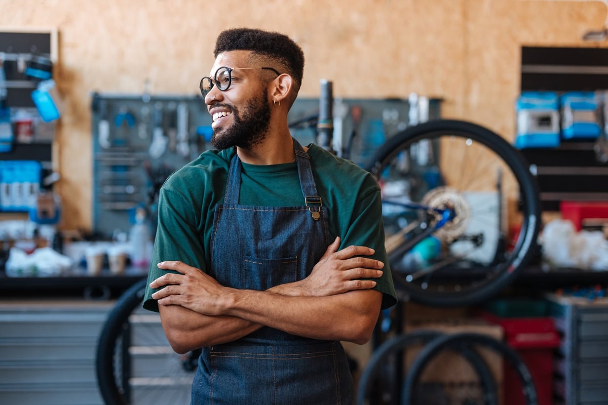 Young African American bicycle mechanic smiling confidently with crossed arms while surrounded by tools and equipment in a bustling workshop