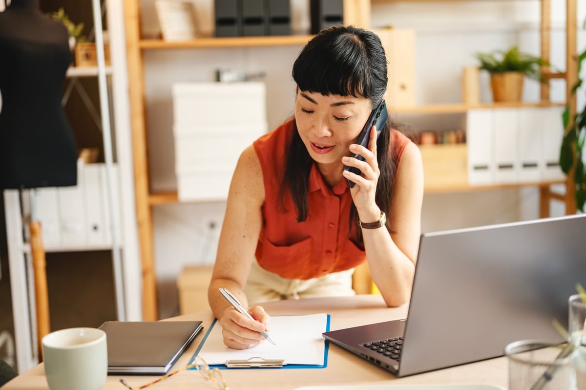 Small business entrepreneur in a modern studio office talking on phone and writing notes at desk with laptop