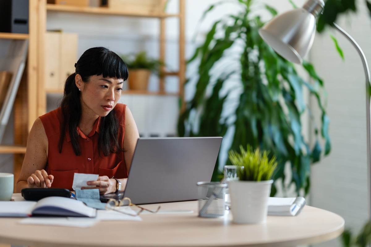 Asian woman entrepreneur in her home office, checking receipts and entering numbers on her phone while working with a laptop and calculator at a round desk