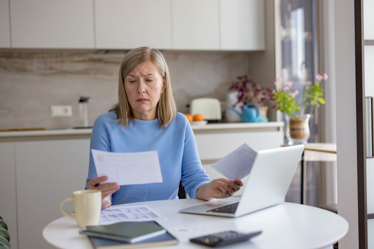 Senior woman at home kitchen table feeling stressed and confused while looking at multiple bills and financial documents