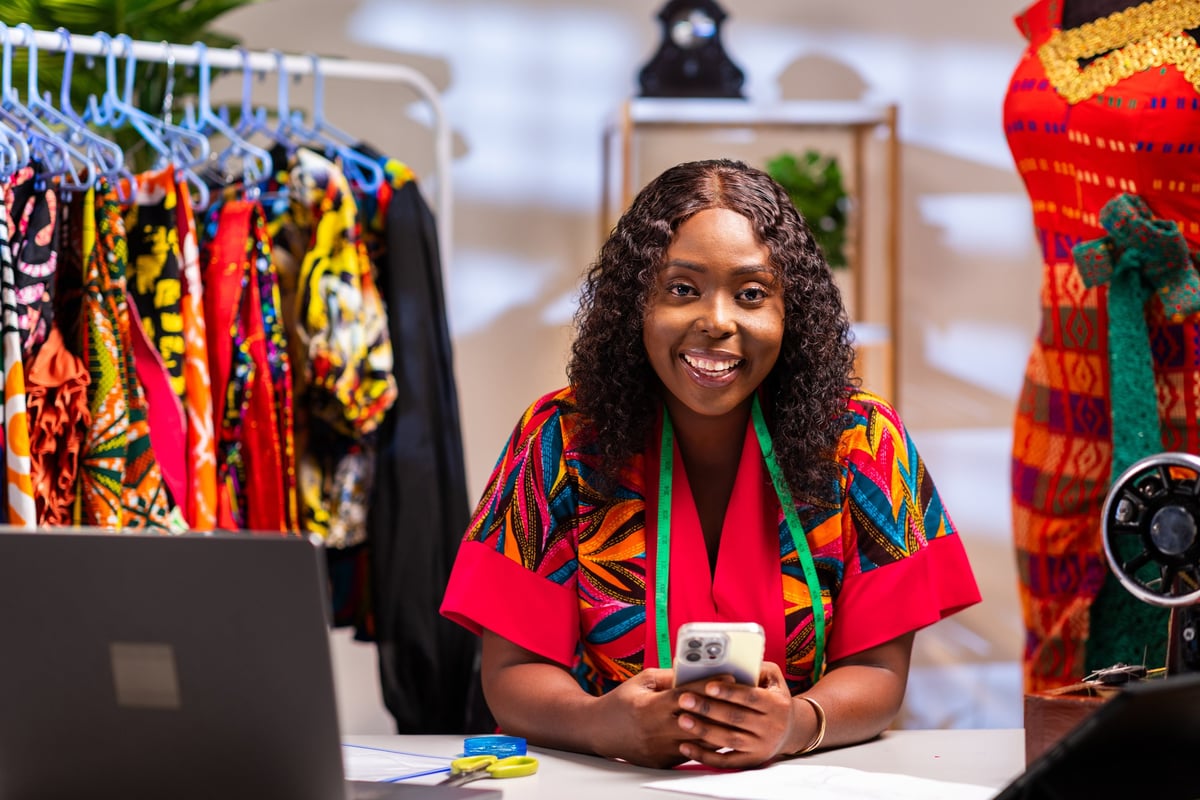 African female fashion design entrepreneur tailor smiling while using her smartphone in her clothing workshop