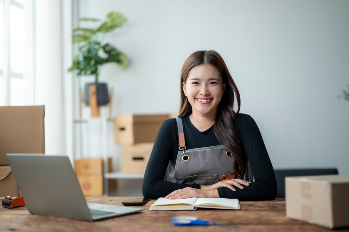 A cheerful woman sits at a wooden desk in a bright office, smiling as she engages with her laptop and notepad