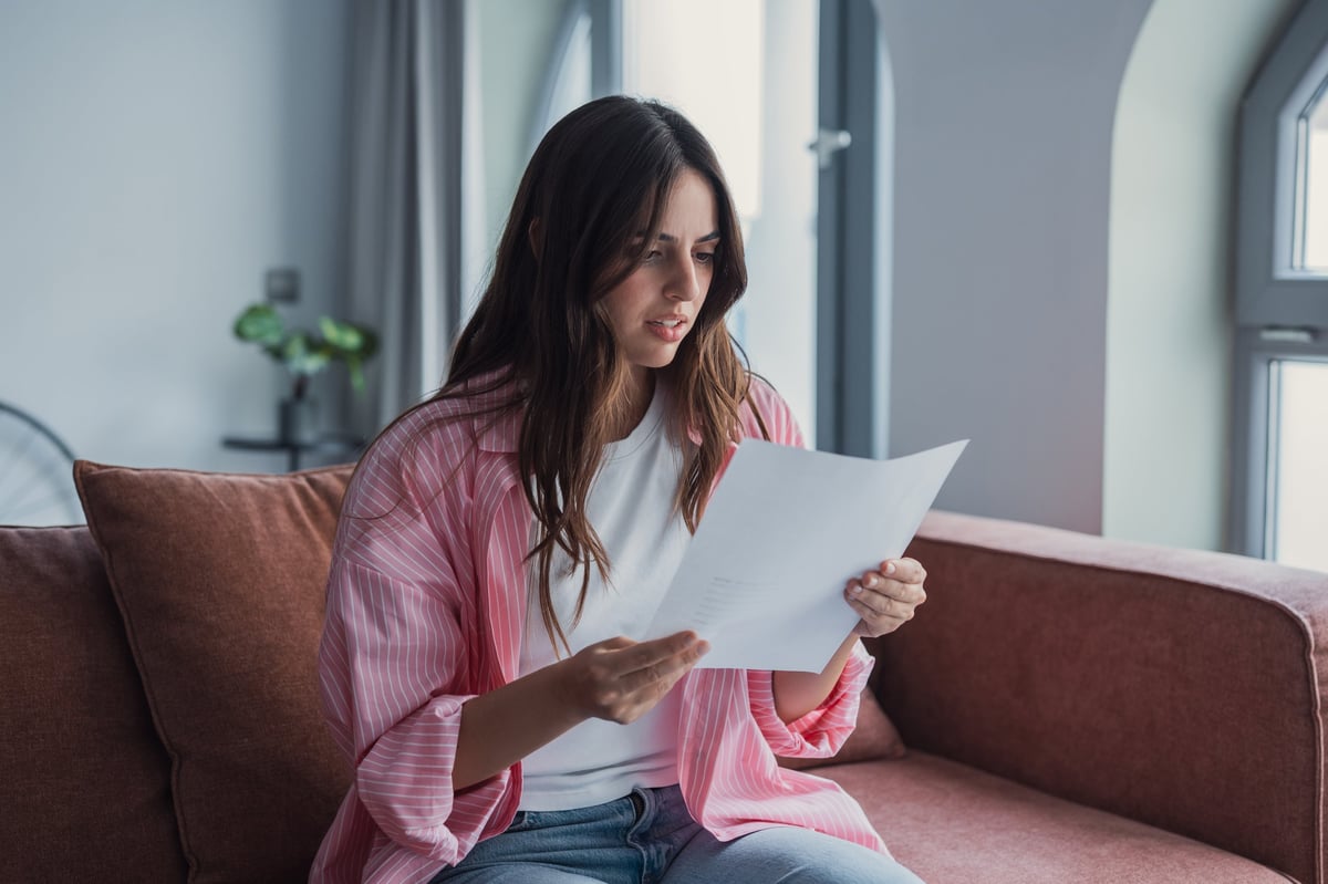 Young woman on a couch in a modern living room reading an important paper or letter with a concerned, thoughtful expression