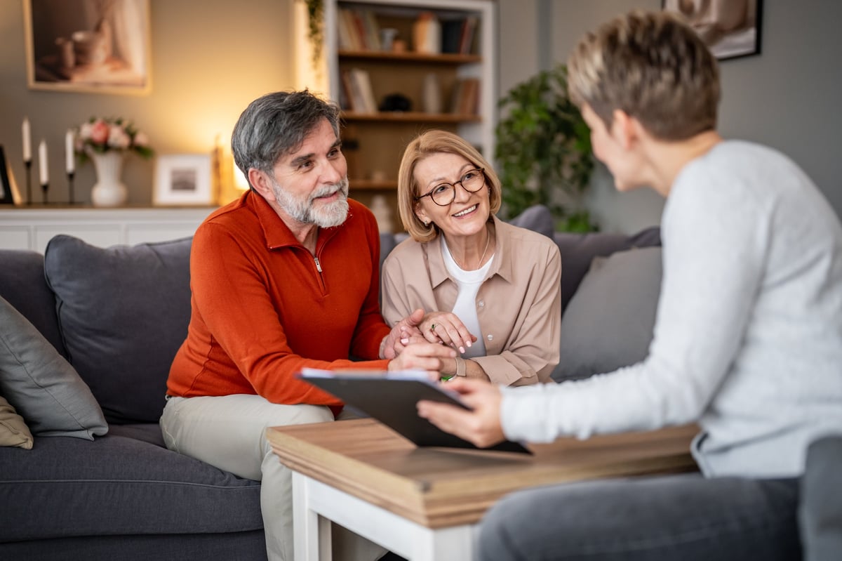 A couple sits on a sofa in a living room, holding hands and talking with an advisor about their home equity options