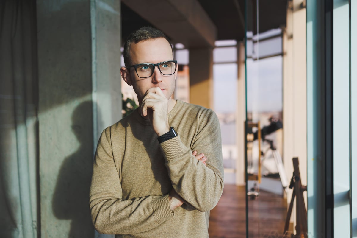 A young man in glasses and casual clothes stands in a modern workspace, male freelancer works remotely