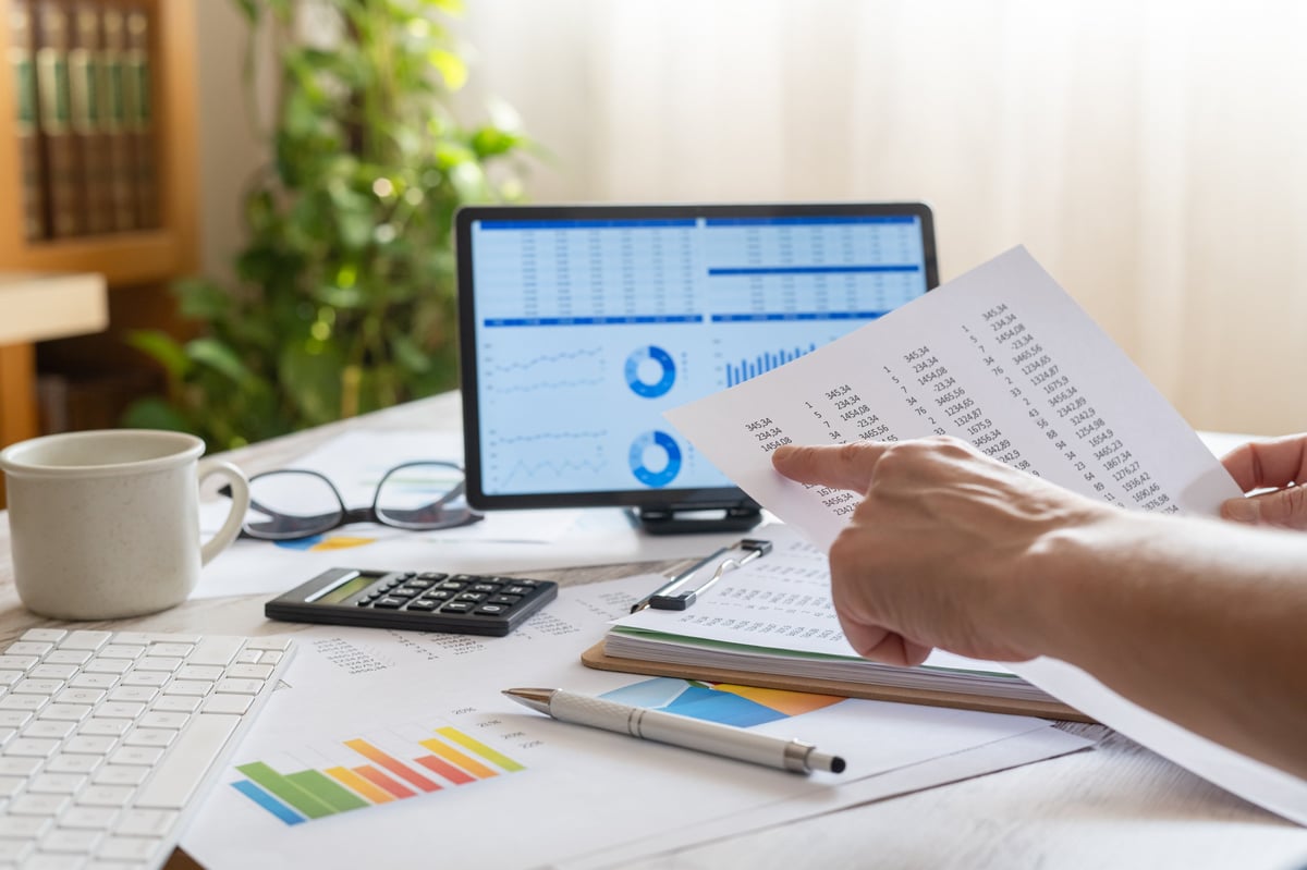 Person's hands closely reviewing a financial report document, pointing at numbers while a laptop displays business charts and graphs on a desk, representing accounting and budgeting tasks