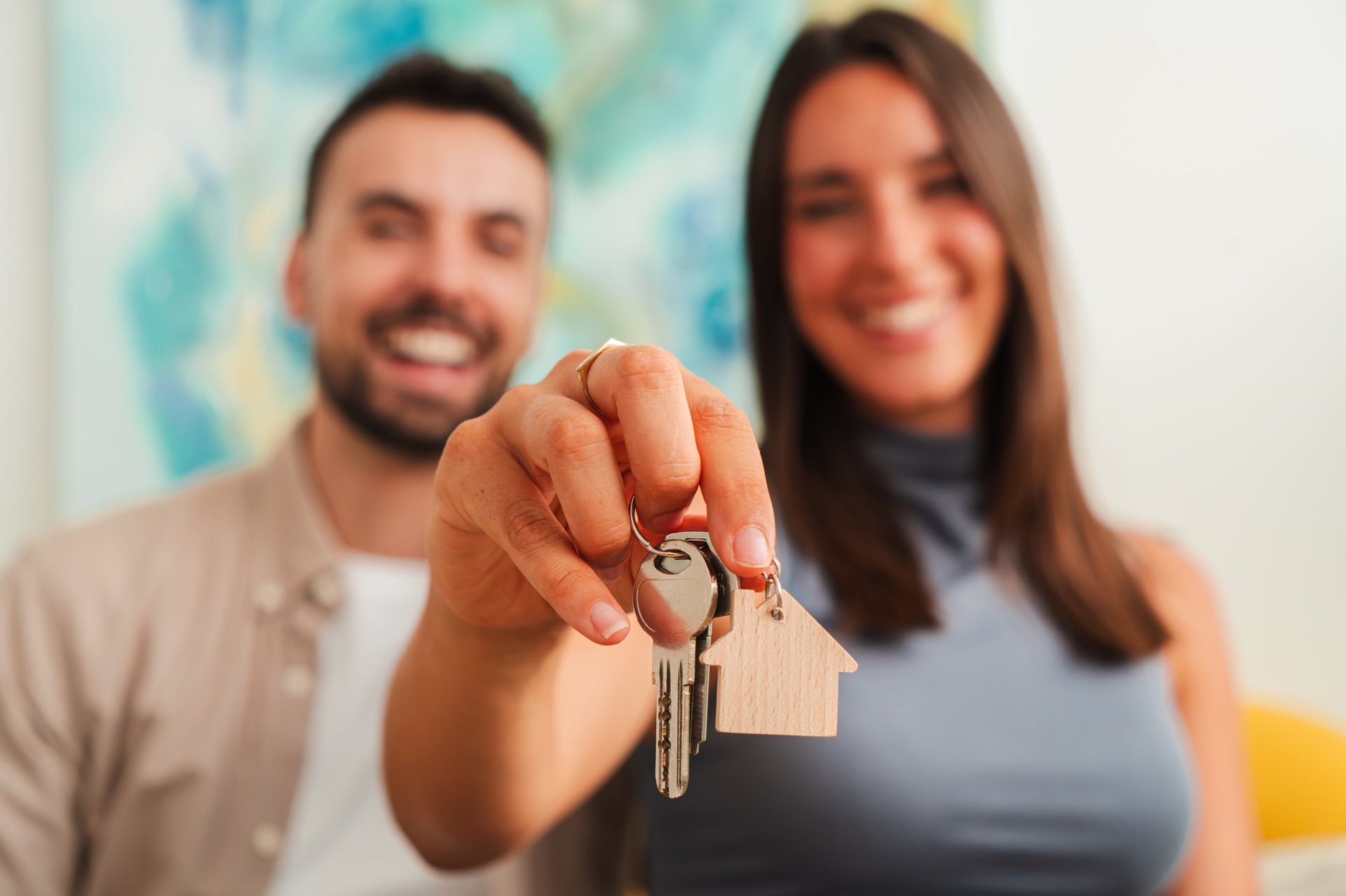 A cheerful couple presents a pair of house keys and a small wooden keychain while standing close, creating a hopeful moment of home ownership and shared excitement