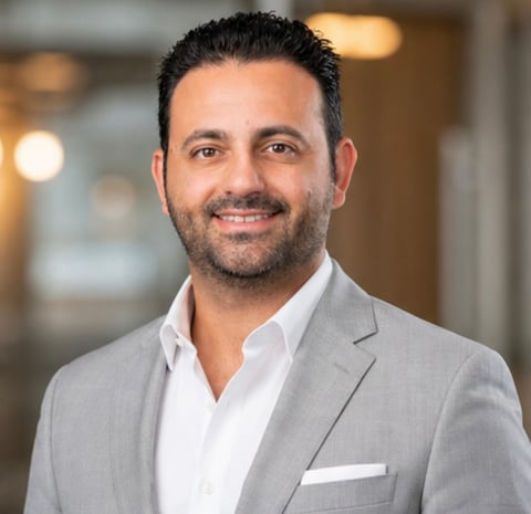 Professional headshot of a man in a light gray blazer and white shirt, smiling at the camera in an office setting
