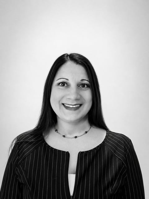 Black and white headshot of a woman with long dark hair, smiling at the camera, wearing a pinstriped blazer and beaded necklace