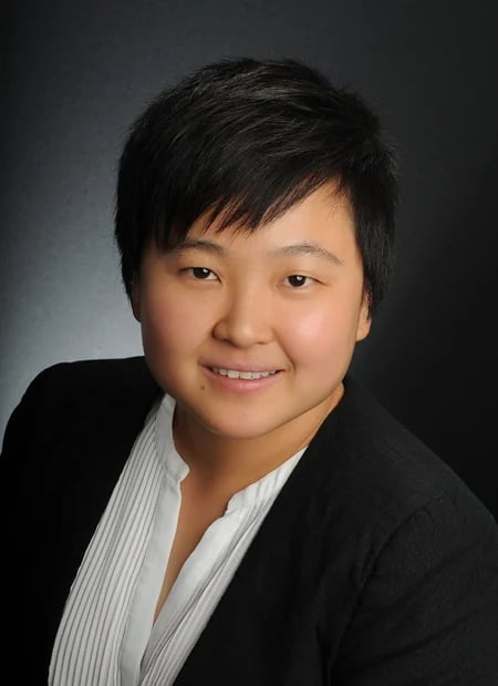 Professional headshot of an Asian woman wearing a black blazer and white striped shirt, smiling at the camera against a gray background