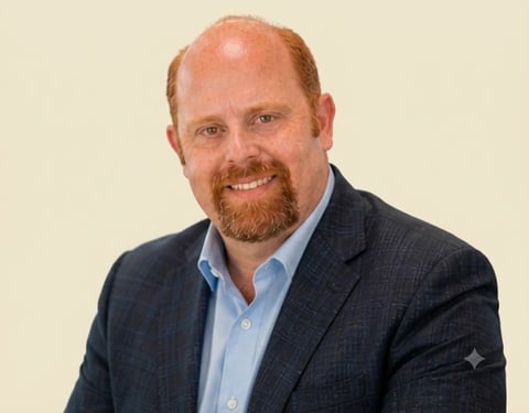 Professional headshot of a man with reddish-brown hair and beard wearing a dark blazer and light blue shirt against a neutral beige background