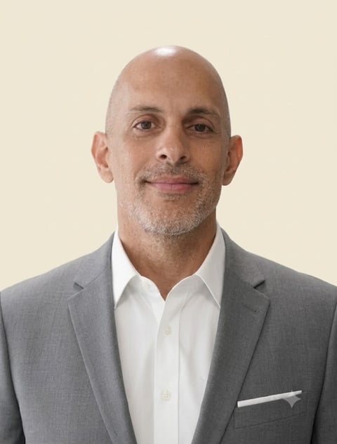 Professional headshot of a bald man in a gray blazer and white shirt against a beige background