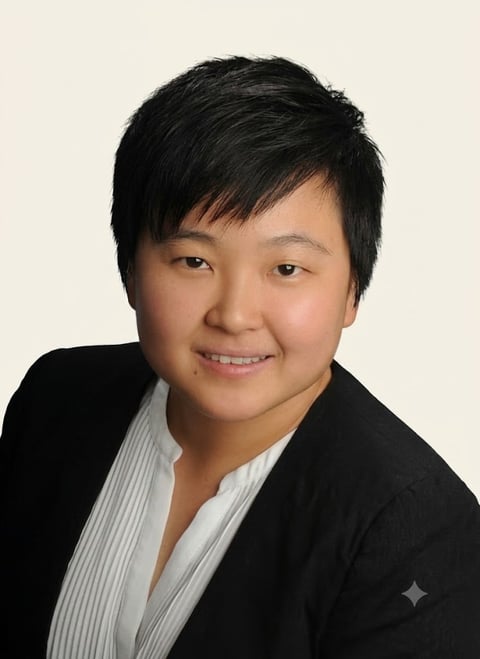 Professional headshot of an Asian person with black hair wearing a black blazer and white striped shirt against a beige background