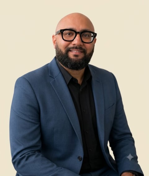 Professional headshot of a man wearing a blue blazer and black shirt, with dark-framed glasses and a full beard, against a beige background.