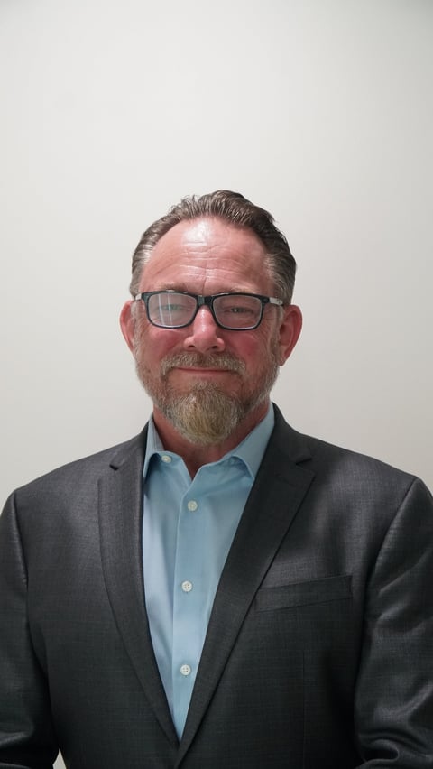 Middle-aged man wearing glasses, dark blazer, and light blue shirt posed against a white background for a professional headshot