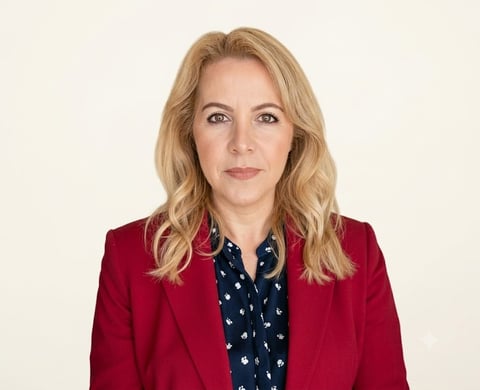 Professional headshot of a woman with blonde wavy hair wearing a red blazer over a navy patterned blouse against a white background