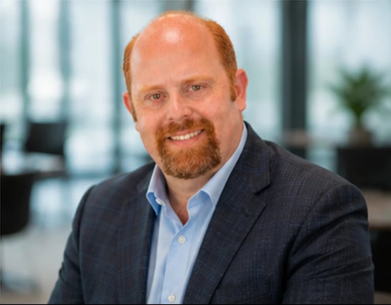 Professional headshot of a smiling man with reddish-brown beard wearing a dark blazer and light blue shirt in a modern office setting