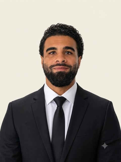 Professional headshot of a man with dark curly hair and beard wearing a black suit and black tie against a cream background