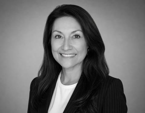 Black and white professional headshot of a woman with dark long hair wearing a dark blazer and white shirt, smiling at the camera