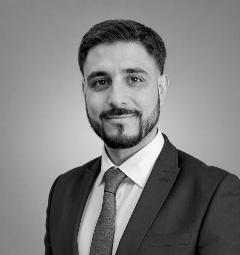 Professional black and white headshot of a man with dark hair and beard wearing a dark blazer and tie