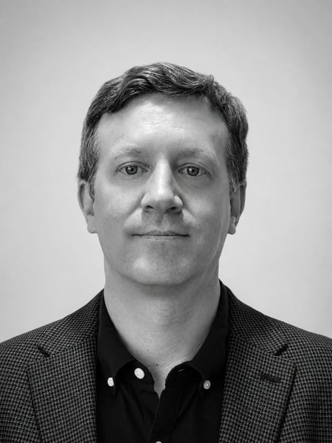Black and white headshot portrait of a middle-aged man wearing a dark blazer and button-up shirt against a plain gray background