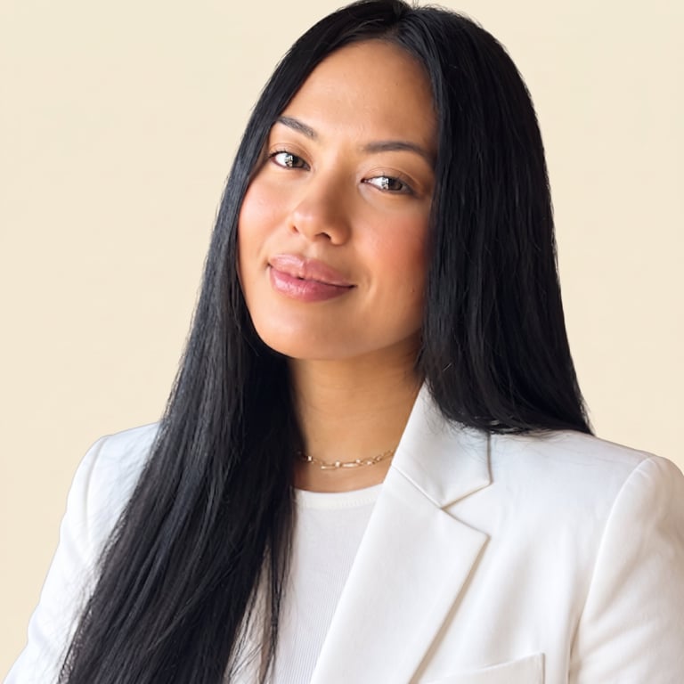 Professional portrait of a woman with long black hair wearing a white blazer and gold chain necklace against a beige background