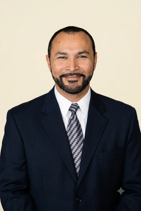 Professional headshot of a man in a dark suit with patterned tie and light blue shirt against a neutral beige background.