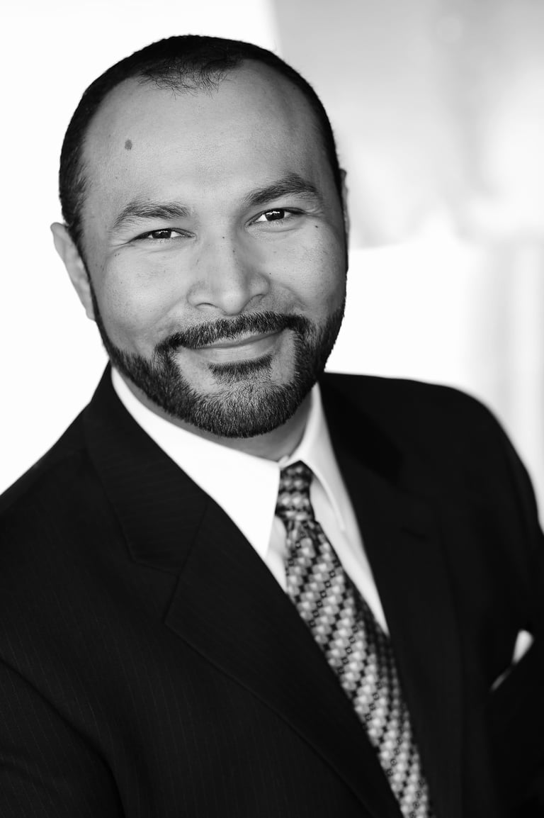 Professional headshot of a man with beard wearing black suit and patterned tie in black and white