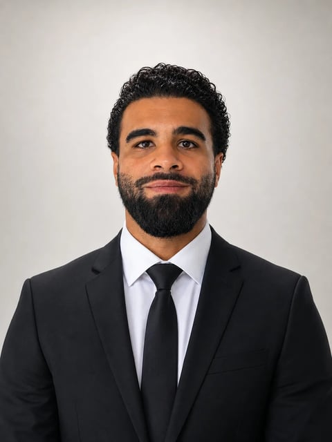 Professional headshot of a man with dark curly hair and full beard wearing a black suit, white dress shirt, and black tie against a white background
