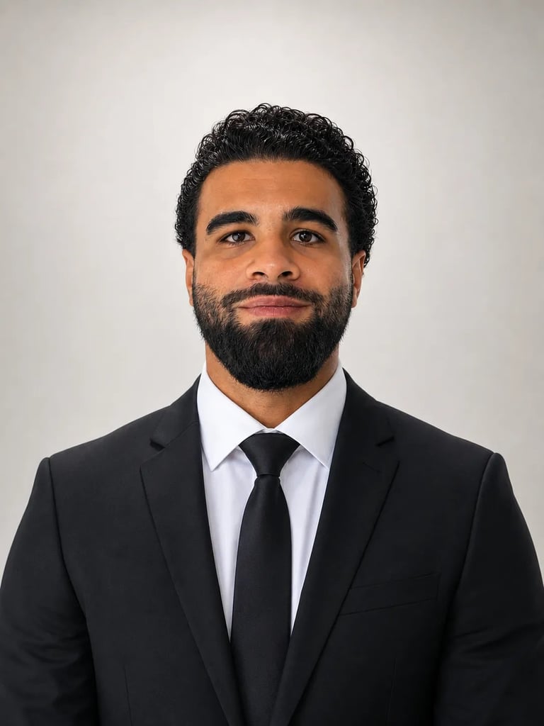 Professional headshot of a man with dark curly hair and full beard wearing a black suit, white dress shirt, and black tie against a white background