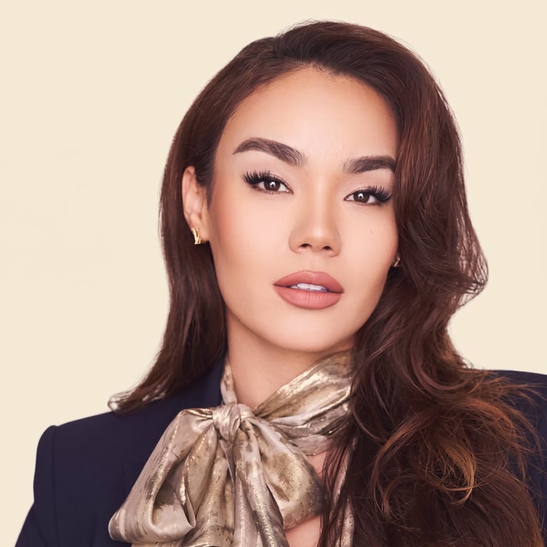 Portrait of a woman with long dark hair and pink lipstick wearing a black blazer and gold silk scarf against a cream background