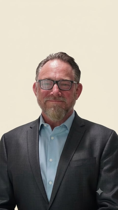 Professional headshot of a middle-aged man wearing glasses, dark blazer, and light blue button-up shirt against a neutral beige background